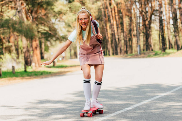 full length view of excited blonde girl skateboarding and listening music in headphones - Valokuva, kuva