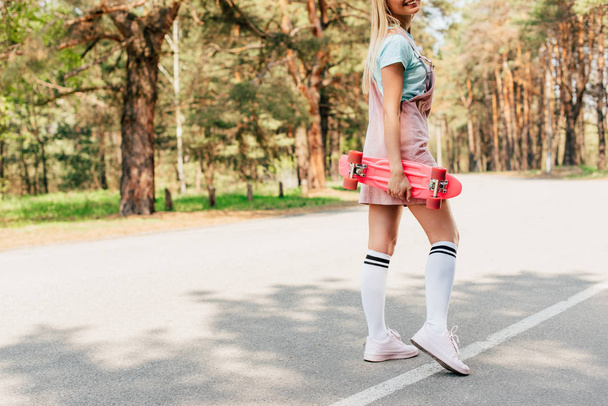 cropped view of smiling blonde girl standing on road and holding penny board - Fotoğraf, Görsel