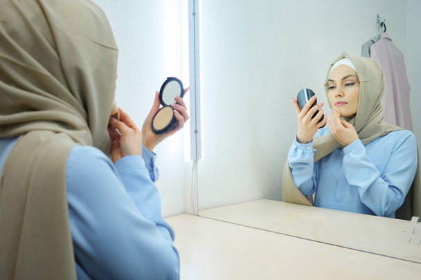 Muslim young attractive woman in beige hijab and traditional blue dress making make-up - Photo, Image