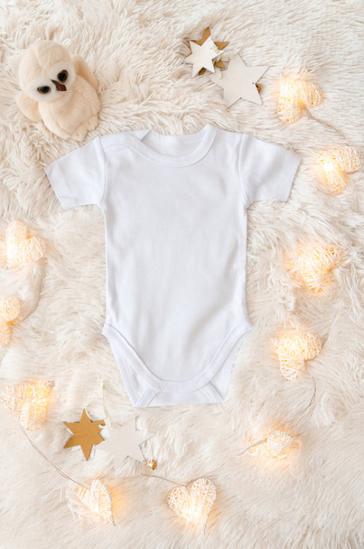 Top view white baby bodysuit on a woolen background. Copy space for text. Accessories and border arrangement with toys and garland of glowing hearts - Foto, Bild