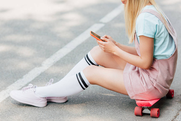 vue recadrée de fille blonde assise sur un panneau de penny et utilisant un smartphone sur la route
 - Photo, image
