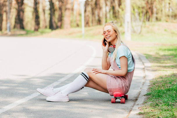 smiling blonde girl in sunglasses sitting on penny board and talking on smartphone on road - Zdjęcie, obraz