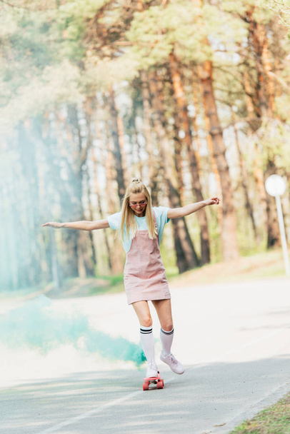full length view of blonde girl waving hands while skateboarding in green smoke on road - Foto, afbeelding