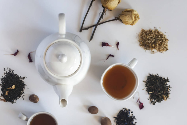Tea set with white ceramic tea pot, dried rose flowers and other tea ingredients on the white. Flat lay view of various dried teas and teapot. View from above. Space for your text - Foto, afbeelding