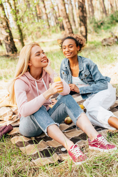  two multiethnic girls sitting on plaid blanket and holding bottles of orange juice - Foto, immagini