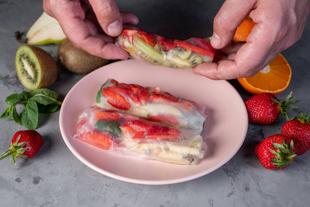 Close view of man holding fruity spring roll over plate with rolls on concrete table - Foto, imagen