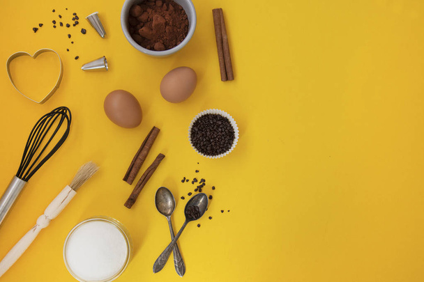 Ingredients for baking with baking utensile on yellow background - flour, wooden spoon, rolling pin, eggs, coccoa, sprinkles, banana. Top view, copy space. - Fotografie, Obrázek