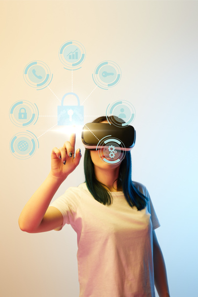 young woman in virtual reality headset pointing with finger at internet security icons on beige and blue background - Photo, Image
