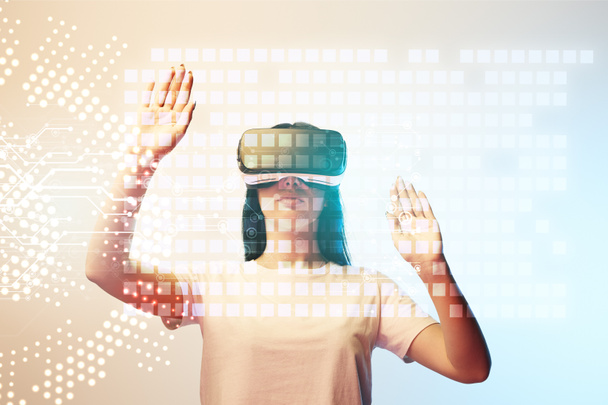 young woman in virtual reality headset pointing with hands at glowing data illustration on beige and blue background - Photo, Image