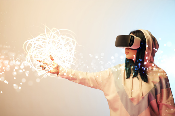 young woman in virtual reality headset pointing with hand at glowing cyber illustration on beige and blue background - Photo, Image