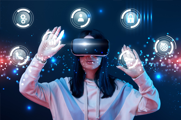 young woman in virtual reality headset gesturing among glowing cyber icons on dark background - Photo, Image