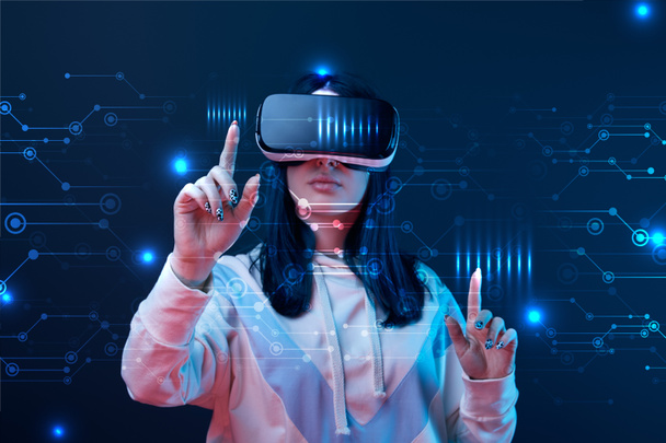 young woman in virtual reality headset pointing with fingers at glowing cyber illustration on dark background - Photo, Image