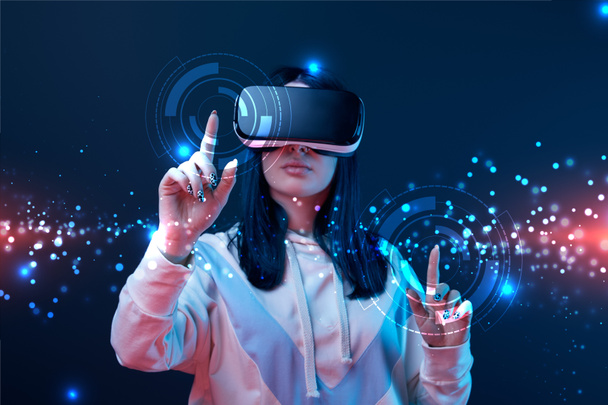 woman in virtual reality headset pointing with fingers at glowing cyber illustration on dark background - Photo, Image