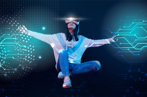 KYIV, UKRAINE - APRIL 5, 2019: Young woman in virtual reality headset with joystick and outstretched hands flying in air among glowing data illustration on dark background  - Fotografie, Obrázek