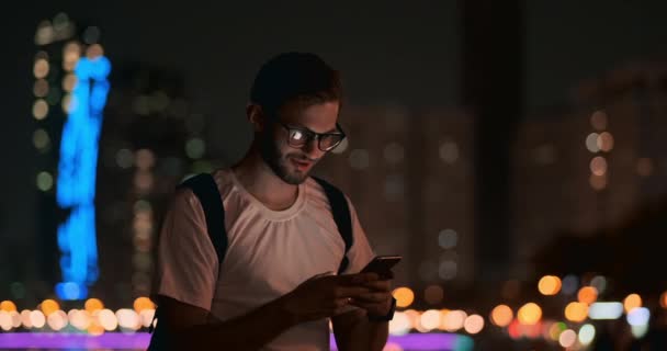 A man in glasses against the background of the night city writes a message using a smartphone - Footage, Video