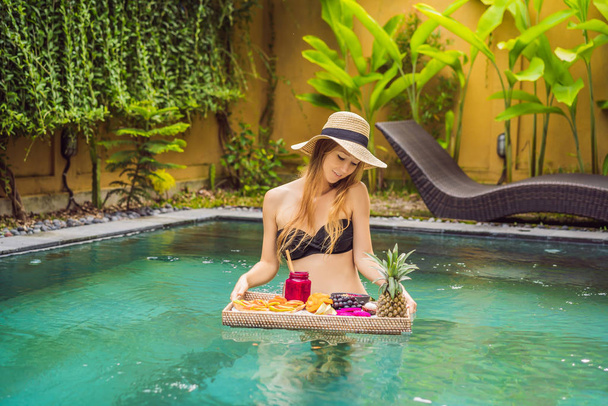 Breakfast tray in swimming pool, floating breakfast in luxury hotel. Girl relaxing in the pool drinking smoothies and eating fruit plate, smoothie bowl by the hotel pool. Exotic summer diet. Tropical - Photo, Image