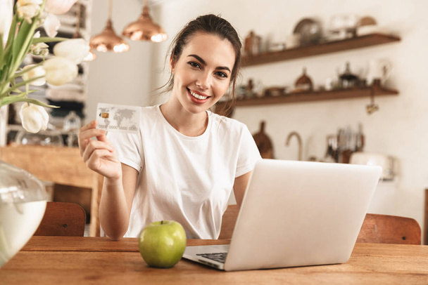 Beautiful young student girl with apple indoors using laptop computer holding credit card. - Foto, imagen
