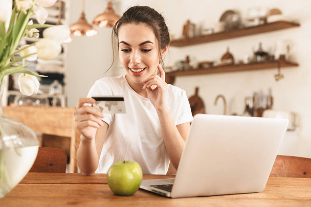 Krásná mladá studentka s jablkem v interiérech používající přenosný počítač držící kreditní kartu. - Fotografie, Obrázek