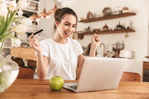 Beautiful young student girl with apple indoors using laptop computer holding credit card. - Фото, изображение
