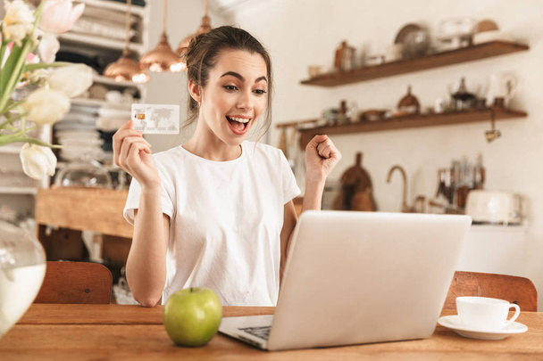 Beautiful young student girl with apple indoors using laptop computer holding credit card. - Foto, Imagem