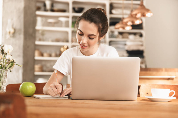 Beautiful young student girl with apple indoors using laptop computer. - Fotó, kép