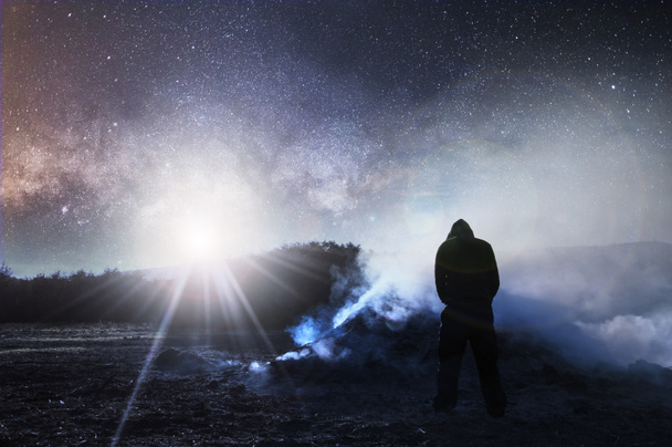 Una edición nocturna de ciencia ficción. Con una figura encapuchada mirando una hoguera y humo con el cielo lleno de estrellas y una luz brillante en el cielo
. - Foto, Imagen