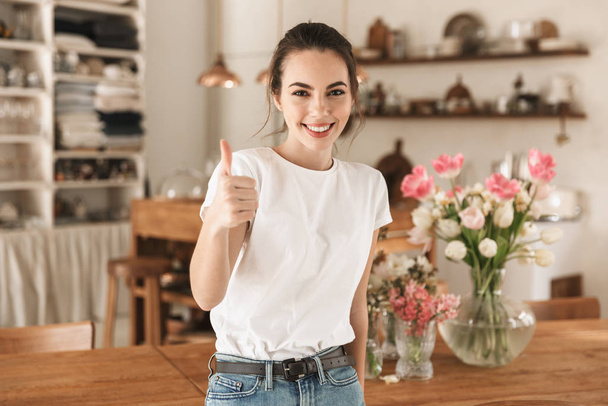 Beautiful young student girl in white t-shirt indoors posing showing thumbs up gesture. - Foto, imagen