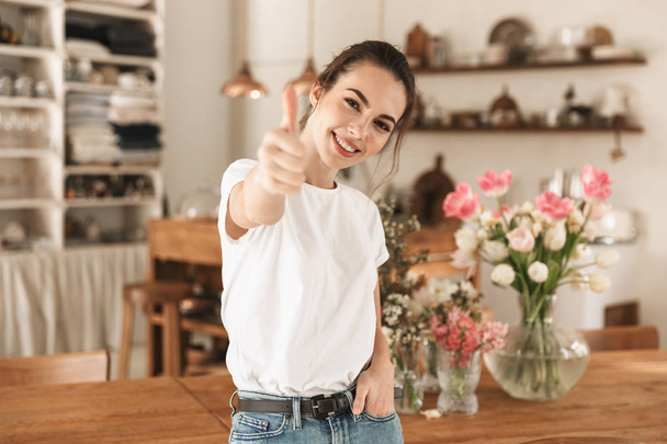 Beautiful young student girl in white t-shirt indoors posing showing thumbs up gesture. - 写真・画像