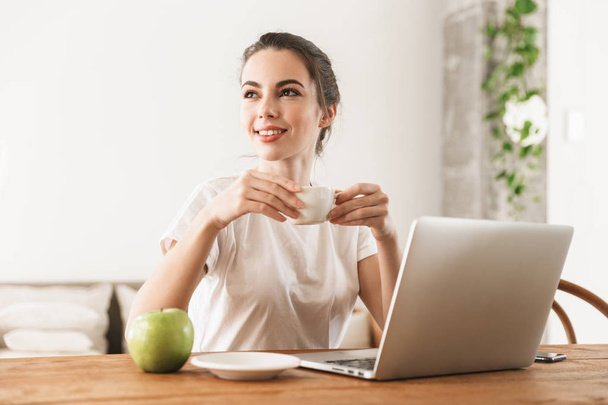 Beautiful young student girl with apple sitting indoors using laptop computer drinking coffee. - Foto, Bild
