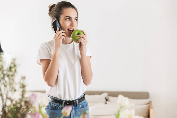 Happy beautiful young student girl eat apple posing indoors talking by mobile phone. - Φωτογραφία, εικόνα