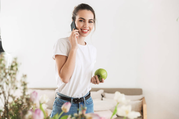 Happy beautiful young student girl eat apple posing indoors talking by mobile phone. - Foto, afbeelding