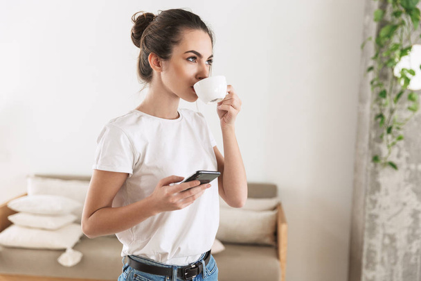 Красивая молодая студентка пьет кофе в помещении, разговаривая по мобильному телефону
. - Фото, изображение