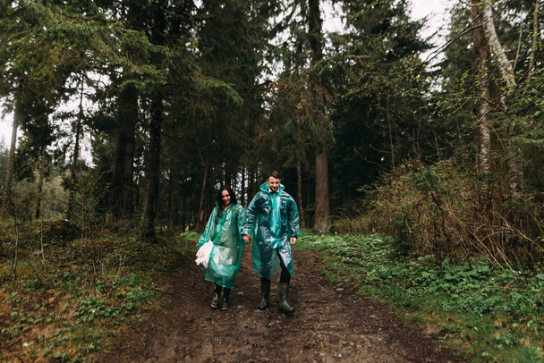 samen alle moeilijkheden overwinnen. Bruid en bruidegom gekleed in regenjassen gaan door het moeras op hun trouwdag - Foto, afbeelding