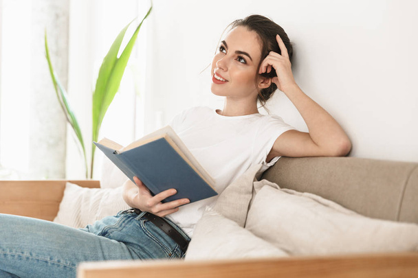Beautiful young student girl sitting indoors reading book on sofa. - Foto, immagini
