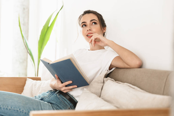 Beautiful young student girl sitting indoors reading book on sofa. - Fotoğraf, Görsel