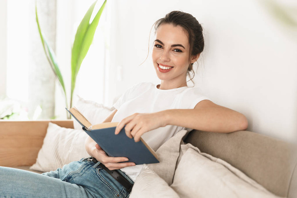 schöne glückliche junge Studentin sitzt drinnen und liest Buch auf dem Sofa. - Foto, Bild