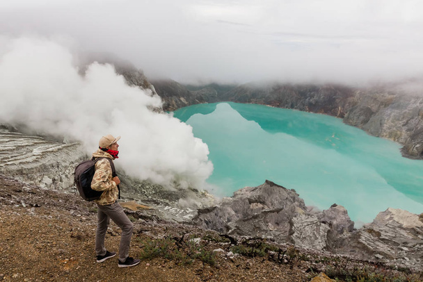Homem turista olha para o lago de enxofre no vulcão Ijen na ilha Java, na Indonésia. Caminhante homem com mochila viajar no topo da montanha, conceito de viagem
 - Foto, Imagem