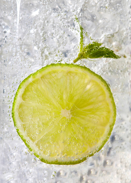 Fresh mint leaf and a slice of lime with bubbles in a glass with ice. Macro photo of refreshing drink - Φωτογραφία, εικόνα
