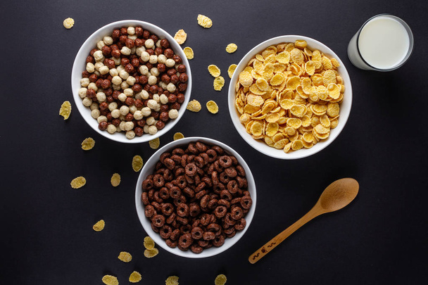 Dry breakfast of chocolate balls, rings and corn flakes. Cereals bowl, spoon and a glass of milk on a black background. Top view  - Foto, Imagem