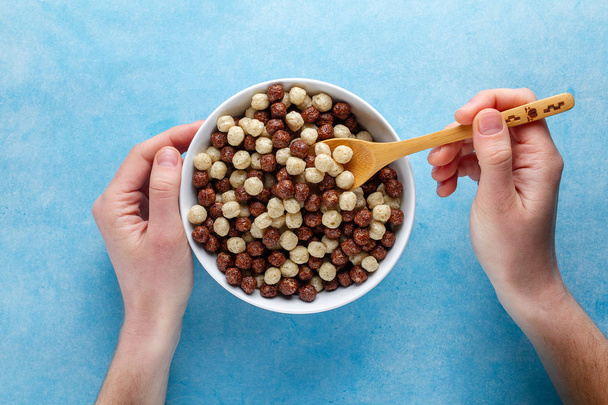 Chocolate, brown and white, glazed balls for dry healthy breakfast. Top view. Cereals spoon and bowl in hands on a blue background  - Photo, image