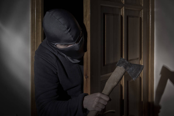 Ladrón con un hacha en la mano y con un sombrero con una hendidura para los ojos se cuela en la casa a través de la puerta por la noche a la luz de la luna
 - Foto, Imagen