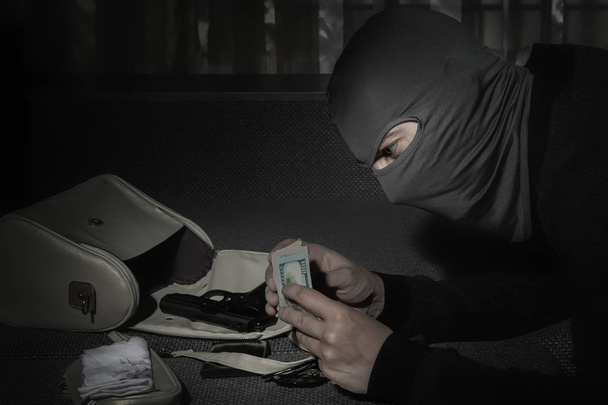 Robber em uma máscara e com uma lanterna examina o conteúdo da bolsa das mulheres em um quarto escuro
 - Foto, Imagem