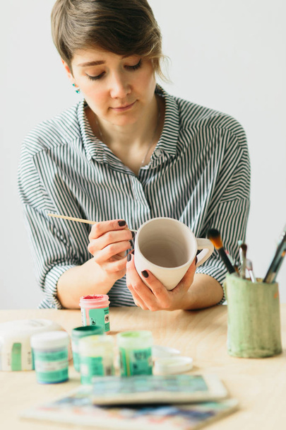 η αγγειοπλαστική γυναίκα κρατά στα χέρια μια κούπα για να ζωγραφίσει το στολίδι - Φωτογραφία, εικόνα
