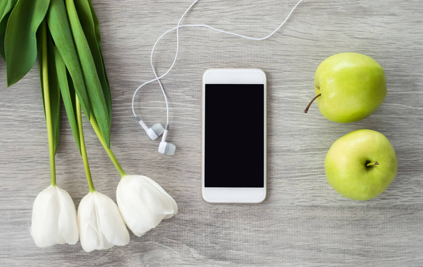 A white phone with white headphones, white tulips and green appl - Foto, Imagen