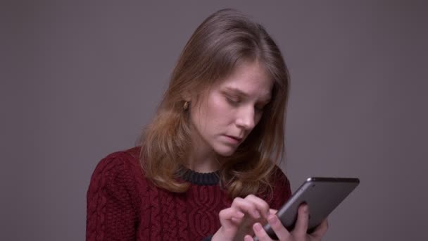 Attentive young female student watching seriously into tablet being concentrated on gray background. - Imágenes, Vídeo