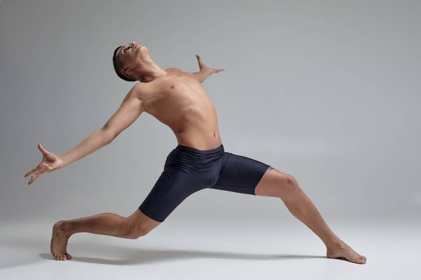 Foto de un hombre guapo bailarín de ballet, vestido con pantalones cortos negros, haciendo un elemento de baile sobre un fondo gris en el estudio
. - Foto, Imagen