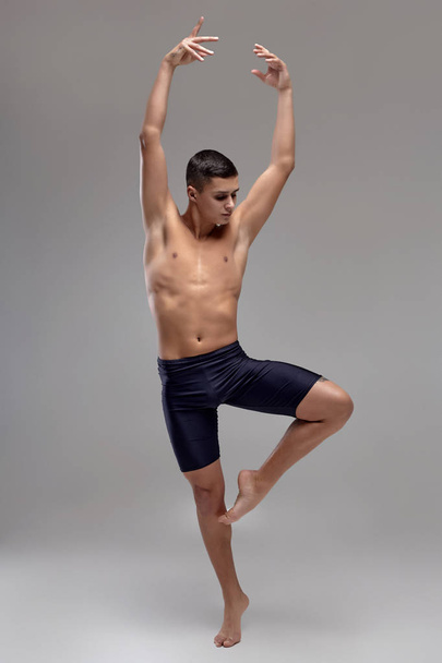Foto van een knappe man ballet danser, gekleed in een zwarte short, het maken van een dans element tegen een grijze achtergrond in Studio. - Foto, afbeelding