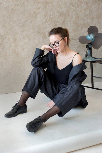 мечтательная девушка в очках, куртка на теле, сидит на полу рядом с фанатом ретро-стиля
 - Фото, изображение