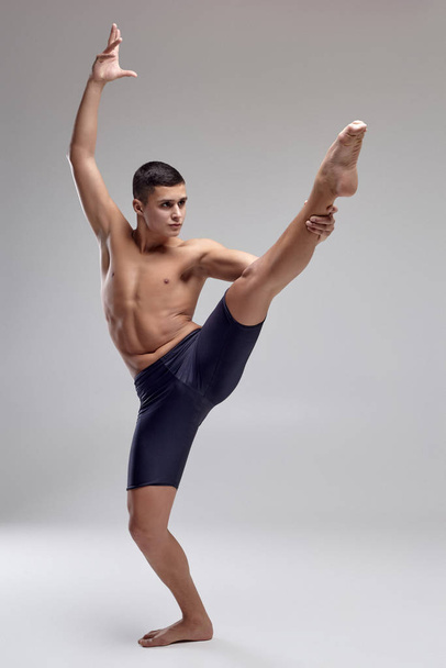 Foto eines gut aussehenden Mannes Balletttänzer, in einer schwarzen Shorts gekleidet, macht ein tänzerisches Element vor einem grauen Hintergrund im Studio. - Foto, Bild