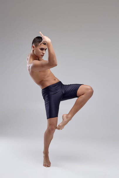 Foto van een knappe man ballet danser, gekleed in een zwarte short, het maken van een dans element tegen een grijze achtergrond in Studio. - Foto, afbeelding